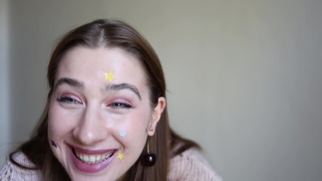Vertical screen young woman presents colorful star shaped acne patches close to camera while wearing one on forehead showing playful skincare and modern blemish treatment