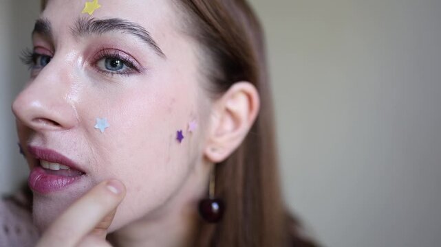 Vertical screen young woman holds colorful star shaped acne patches close to camera while wearing stickers on her face presenting playful skincare and modern blemish care