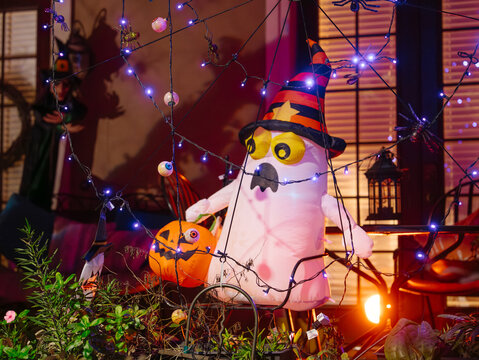 A large inflatable ghost in a witch hat stands beside an orange jack-o-lantern. Glowing purple lights, spiders, and eyeballs decorate the residential porch for Halloween.