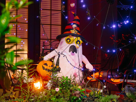 A white inflatable ghost holds a pumpkin while standing among garden plants on a porch. Purple spiderweb lights, spiders, and hanging eyeballs create a spooky Halloween scene.