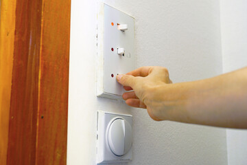Naklejka premium Hand flips lower toggle on triple light switch panel with red indicators on white wall. Shows household grid load reduction, conscious energy conservation habits, and residential utility management.