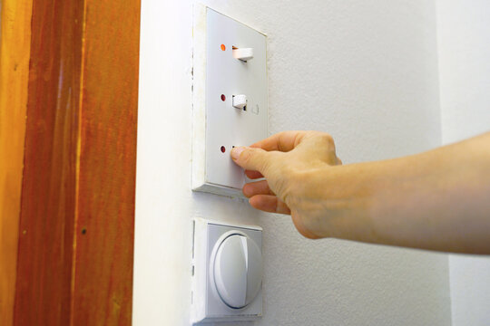 Hand flips lower toggle on triple light switch panel with red indicators on white wall. Shows household grid load reduction, conscious energy conservation habits, and residential utility management.