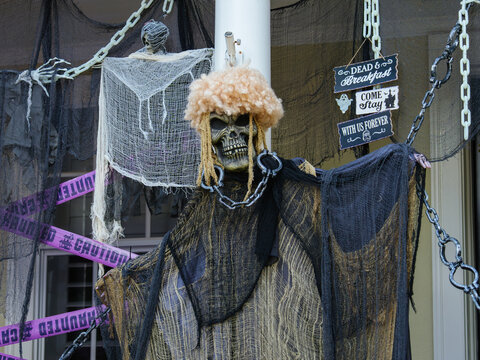A life-size skeleton figure stands among spooky decorations, chains, and warning signs on a haunted Halloween porch. The dramatic display creates an eerie seasonal scene.