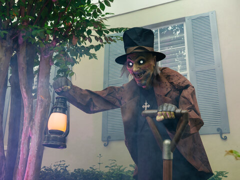 A spooky Halloween figure with glowing eyes holds a lantern and shovel in a foggy front yard. The haunted decoration creates a chilling atmosphere outside a residential home.