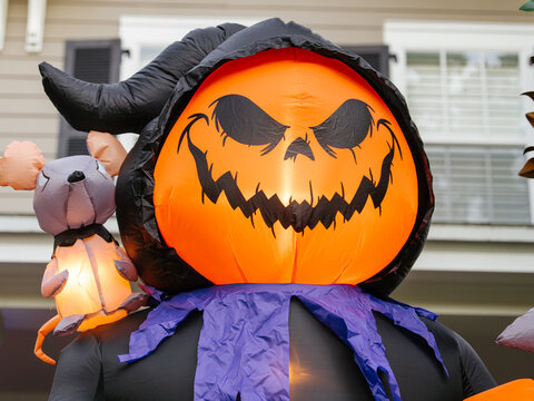 A giant inflatable Halloween reaper with a glowing pumpkin face stands outside a suburban home. The illuminated decoration adds a fun and spooky touch to the holiday display.