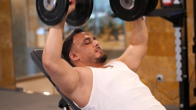 Muscular athletic man lying on incline bench performing heavy dumbbell chest press in a modern indoor fitness club