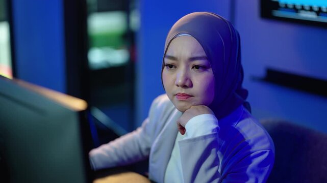 Tired Muslim businesswoman in hijab working late at night in a dark office illuminated by blue computer screen light. Stressed female employee focuses on problem solving, looking surprised.