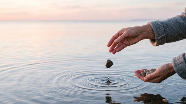Person dropping a small stone into the calm water of a lake or ocean creating circular ripples during a peaceful sunset