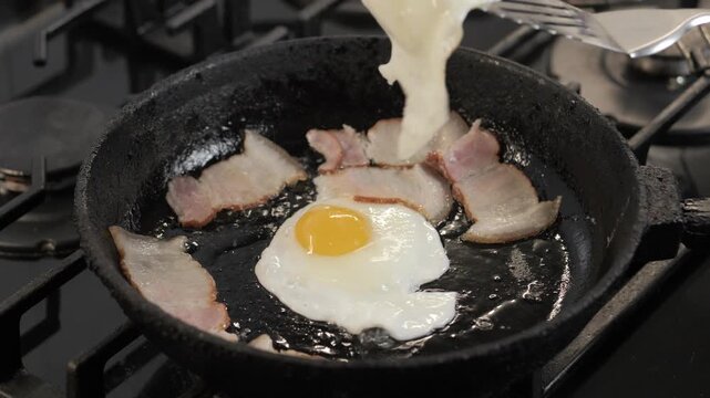 Slotted spatula lifting and flipping fried eggs over bacon in cast-iron skillet on gas stove, with egg, yolk, spatula, bacon detail, showing skillet, castiron, stove, pan context