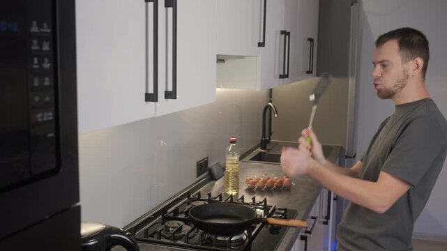 Man preparing breakfast in modern kitchen handling spatula over frying pan on gas cooktop with eggs and oil on countertop, with kitchen, cooktop, stove, pan detail