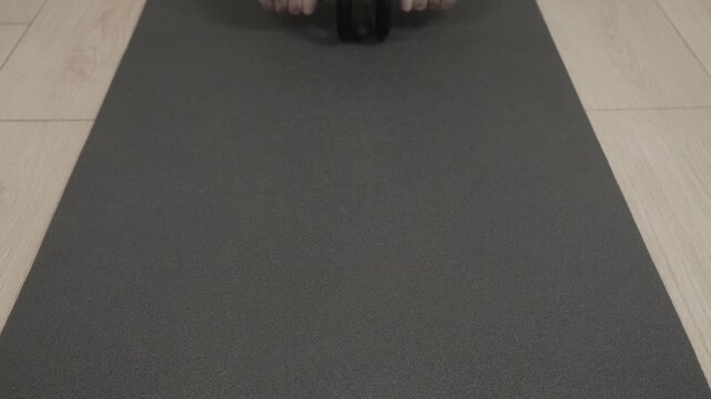Male adult kneeling on black exercise mat performing ab roller rollout toward camera, hands gripping dual-wheel roller while torso leans forward and head tilts down showing concentrated effort