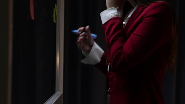 Businesswoman brainstorming ideas writing on glass board. Pensive professional woman writing on a sticky note and putting it on a glass board. Project manager developing a new business strategy.