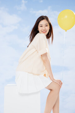 Smiling young woman sitting with yellow balloon against blue sky