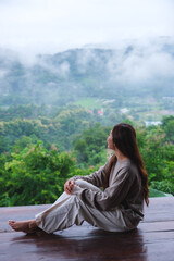 Naklejka premium Vertical image of a woman sitting on wooden balcony with a beautiful mountain views on cloudy morning