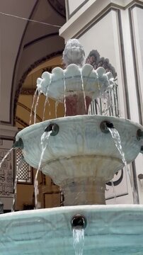 Close-up view of a traditional ornate marble fountain with multiple tiers and cascading streams of water. Decorative Islamic architectural elements and Arabic calligraphy are visible in the background