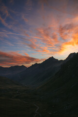 Obraz premium Dramatischer Sonnenuntergang über einem alpinen Bergtal mit kurvenreicher Bergstraße und leuchtendem Abendhimmel