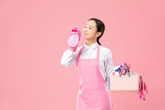Smiling housekeeper with spray bottle and cleaning supplies on pink background