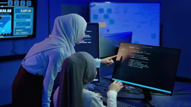 Two Muslim women programmers in hijabs collaborating on software code late at night in a dark high-tech office. Female IT developers discussing cybersecurity data on multiple computer screens.