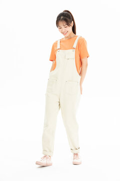 Smiling young woman in light overalls and orange t-shirt standing on white background