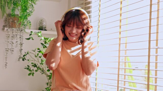 Woman in Sunlight Beside Window Blinds Adjusting Hair, Peaceful Home Interior Portrait with Leafy Plant, Warm Shadow Patterns, Headphones, Smiling Face, and Relaxed Indoor Music Lifestyle Moment