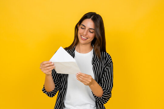 Woman smiling with closed eyes, holding an open envelope with a letter or important message inside, standing against a bright yellow background, conveying happiness and positive news
