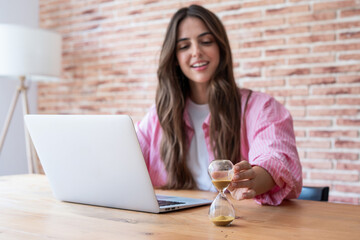 A young woman working at a laptop in a cozy home office turns an hourglass to track time,...