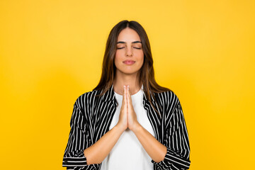 Obraz premium Young woman standing with eyes closed and hands pressed together in a prayer or meditation gesture, finding inner peace, mindfulness, gratitude, and spiritual connection on a yellow background