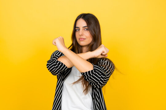 Woman standing with a serious expression, making a refusal or protest gesture by crossing her arms in an x shape against a vibrant yellow background, indicating denial, rejection, or boundaries
