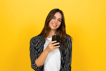Naklejka premium Young woman smiling and holding a smartphone, enjoying social media and messaging on a vibrant yellow background, casual portrait conveying joy, connectivity, and modern lifestyle