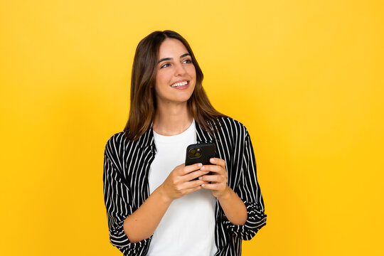 Smiling young woman holds a smartphone and gazes away against a vibrant yellow background, communicating a sense of happiness, connection, optimism, casual style and modern digital lifestyle.