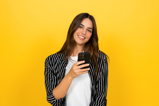 Young woman smiling and holding a smartphone, enjoying social media and messaging on a vibrant yellow background, casual portrait conveying joy, connectivity, and modern lifestyle