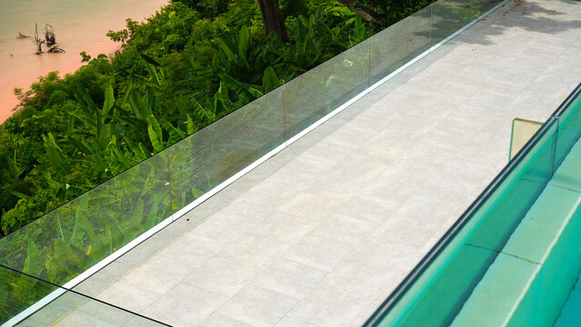 High-angle view of a modern tiled balcony with a sleek transparent glass railing overlooking a vibrant green tropical forest and a muddy river below