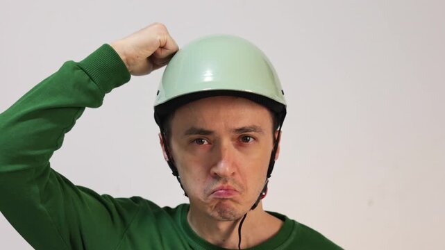 Comical man wearing a safety helmet making funny faces and repeatedly punching himself on the head with his fist, isolated on a white background. The concept of stupidity and odd behavior