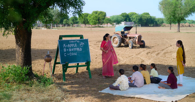 Indian happy mature adult gen x female hold stick do work talk to villager girl boy sitting on mat rural area teen male child study look board enjoy desi life open day class field ground place