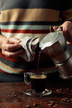 A man in a striped sweater is poured coffee from a geyser coffee maker into a glass cup.