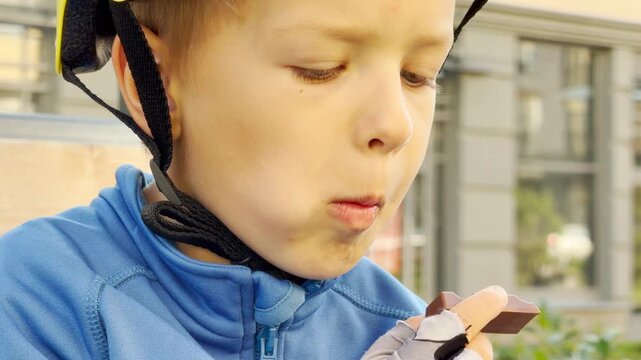 A young boy in a helmet eats chocolate outside. He happily enjoys the sweet treat in a bright urban street. The content child feels satisfied and pleased.