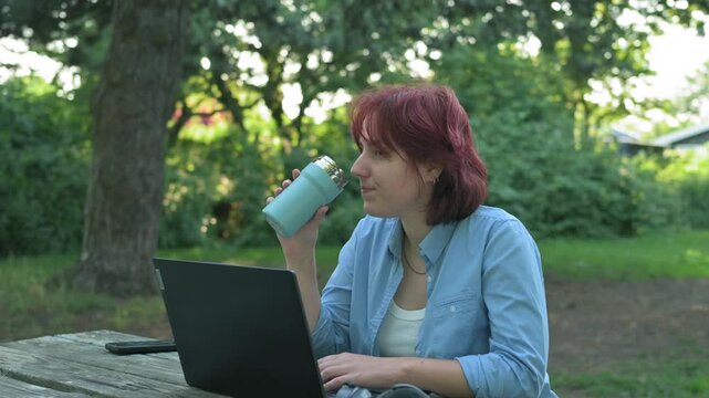 Red-haired woman sipping coffee from reusable eco thermal cup in park.