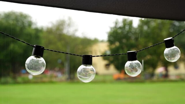 Close-up video of decorative string light bulbs hanging under a garden umbrella during a rainy summer day. Blurred green park background with gentle raindrops falling.