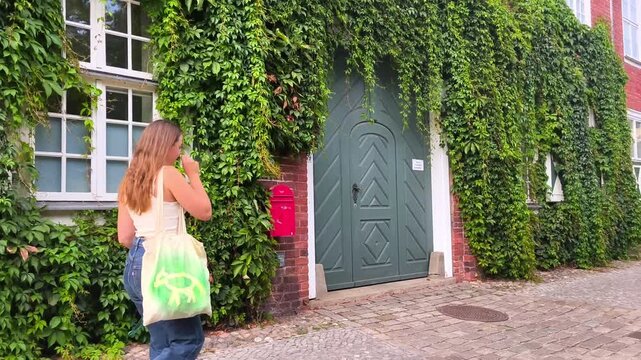 Potsdam, Germany - August 29, 2026: Young woman approaches a green door covered in ivy, carrying a reusable bag, with a brick wall and windows visible in the background