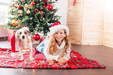 Naklejka premium Girl in santa hat lying on plaid blanket with dog near decorated christmas tree with gifts. Child and pet celebrating winter holidays at home. Family celebration concept