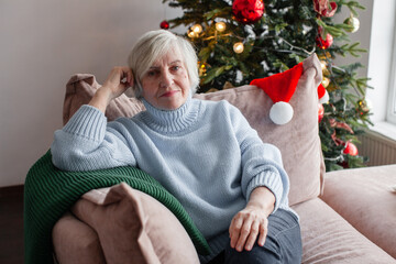 Naklejka premium Senior woman relaxing on couch at home near decorated Christmas tree. Elderly woman celebrating winter holidays with Santa hat. Cozy Christmas celebration concept.
