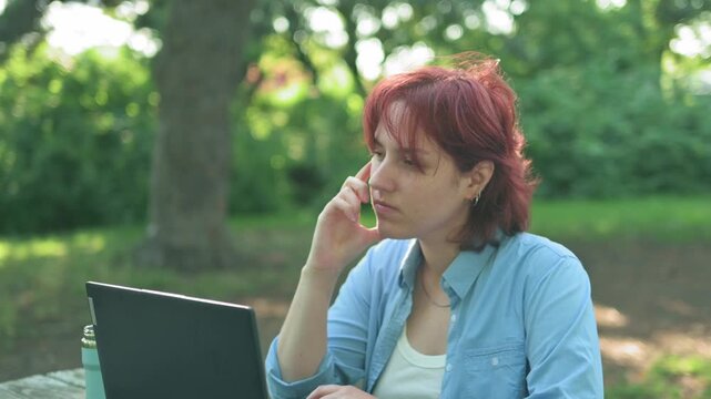 Thoughtful redhead woman looking away in search of inspiration then typing on laptop in the park.