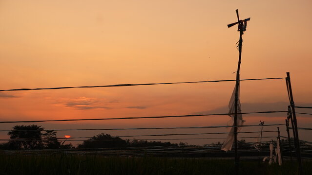 A low-angle perspective captures the orange sun setting near the horizon, behind unfurled plastic ribbons and a wooden turbine to keep birds away