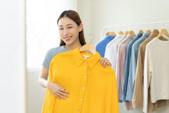 Choose clothes to wear, attractive asian young woman, girl try on appare, happy choice of shirt, female outfit on hanger in wardrobe in room closet at home. Deciding blouse what to put on which one