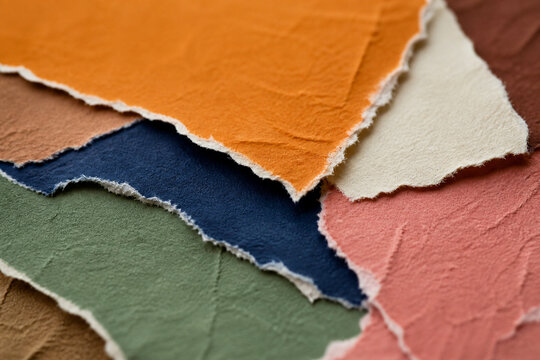 Close-up of overlapping handmade paper sheets in warm and muted colors, showing rich texture and torn deckled edges.