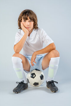 Young soccer player with long hair, sitting on soccer ball with face resting on his hand casually