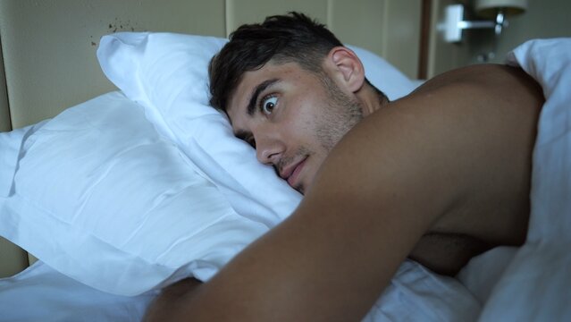 Young man suddenly waking up in bed with surprised expression in modern hotel room or home interior. Emotional guy reacting to abrupt awakening showing sleep interruption and natural human reaction