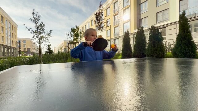 A focused young boy plays table tennis as an orange ball bounces near his paddle. He stands in a sunny courtyard near modern buildings. His happy smile shows pure excitement and joy.