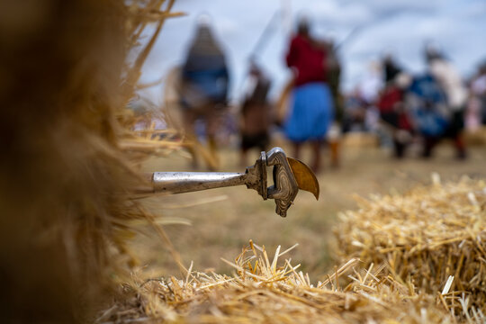 Forged medieval battle axe head stuck in a straw bale with blurred reenactors fighting in historical battle on the background.
