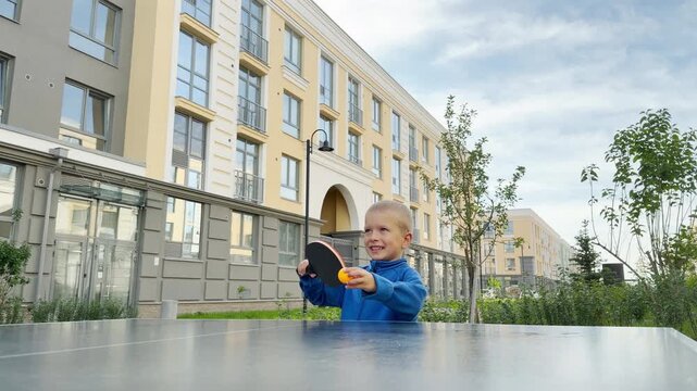 A happy young boy holds a paddle and orange ball to serve. He plays table tennis in an outdoor courtyard near modern buildings. His cheerful smile shows pure joy and excitement.